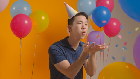 Studio-Portrait-Of-Man-Wearing-Party-Hat-Celebrating-Birthday-Blowing-Paper-Confetti